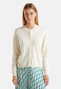 Bílý cardigan vyrobený z texturované pleteniny, s kulatým výstřihem a knoflíky vpředu, nošený přes vzorovanou modro-bílou sukni.