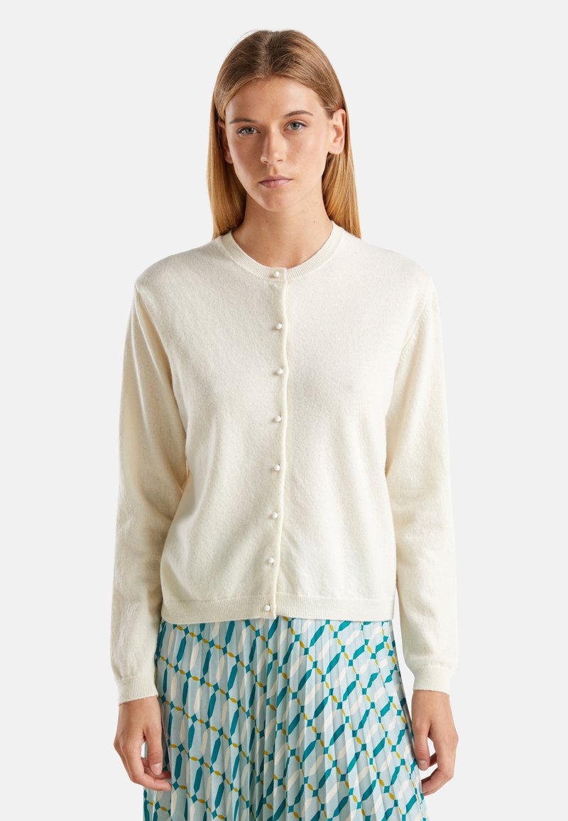 Bílý cardigan vyrobený z texturované pleteniny, s kulatým výstřihem a knoflíky vpředu, nošený přes vzorovanou modro-bílou sukni.