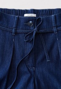 Pantalon en denim bleu foncé avec ceinture élastique, fermeture par bouton, passants pour ceinture, ceinture en tissu nouée et étiquette "OPUS" visible sur la ceinture.