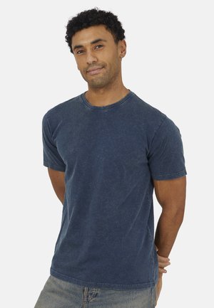 Uomo con capelli ricci che indossa una maglietta a maniche corte blu scuro slavata e jeans chiari, in piedi con le mani dietro la schiena.