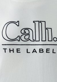 Valge T-särk, millel on musta tikandiga tekst "Calli. THE LABEL." Kangas näeb välja pehme ja sile.