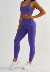 Brassière de sport violette et leggings taille haute faits d'un tissu extensible. Présente un petit logo en forme de cœur sur la ceinture et une texture lisse.