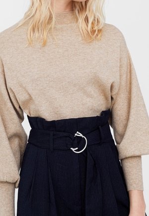 Pull en maille beige à manches longues associé à un pantalon noir à rayures fines taille haute avec une large ceinture ornée d'une boucle circulaire argentée.