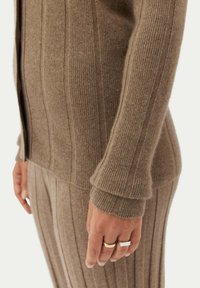 Brun ribbad stickad tröja med vertikala ränder, med långa ärmar och en avslappnad passform. Texturen är mjuk och varm.