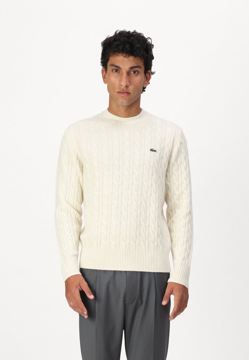 Cream cable-knit sweater with a round neckline, ribbed cuffs and hem. Features a small green logo on the left side of the chest.
