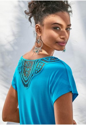 Turquoise short-sleeve top featuring a lace crochet detail on the back neckline. Model is wearing large silver earrings.