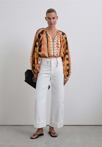 Blusa de múltiples colores con un estampado, escote en V y cordón, combinada con pantalones anchos blancos y un bolso de mano negro. Tejido texturizado y fluido.