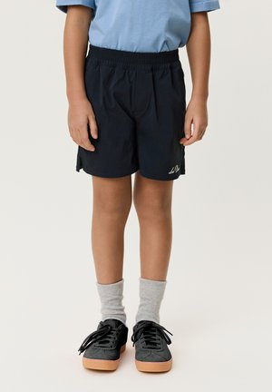 Barn iført sorte elastiske shorts, lyseblå kortærmet skjorte, grå sokker og sorte sneakers med gummisåler, stående på en hvid baggrund.