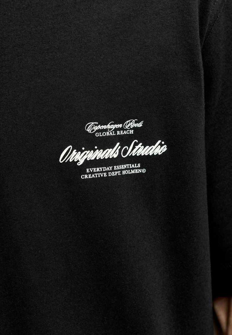 Μαύρο ύφασμα με λευκό κείμενο που λέει Copenhagen Roots Global Reach Originals Studio Everyday Essentials Creative Dept Holmen© στην περιοχή του στήθους.