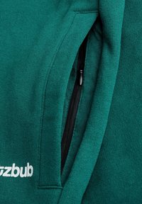 Grüner Sweatshirt mit weicher Textur, ausgestattet mit einer Seitentasche mit schwarzem Reißverschluss und weißem Logotext nahe dem Saum.