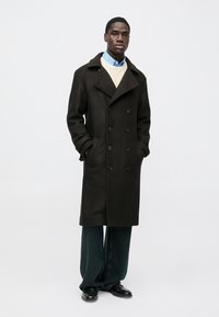 Dark brown wool overcoat with double-breasted design, large collar, and black buttons. Paired with a light sweater, blue shirt, and wide-leg jeans.