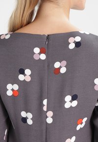 Le tissu gris présente un motif floral avec des pois blancs, roses, rouges et bleu marine. Une petite fermeture éclair est visible au niveau de l'encolure arrière.