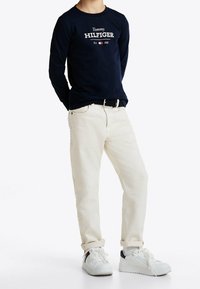 Marineblaue Langarmbluse mit weißem "Tommy HILFIGER"-Text, kombiniert mit hellbeigen Hosen und weißen Sneakers mit marineblauen Akzenten.