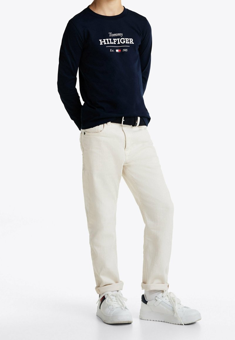 Marineblaue Langarmbluse mit weißem "Tommy HILFIGER"-Text, kombiniert mit hellbeigen Hosen und weißen Sneakers mit marineblauen Akzenten.