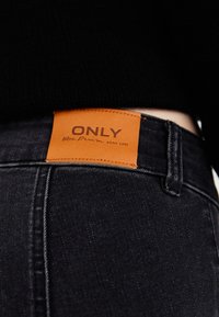Črne kavbojke iz denima z oranžno oznako blagovne znamke, na katero je na pasu všito besedilo "ONLY Blue Denim anno 1995". Gladka tekstura in ozek kroj.