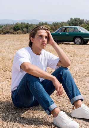 Un jeune homme est assis dans un champ sec, la tête reposant sur une main, avec une voiture ancienne verte garée derrière lui.