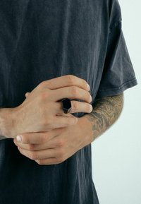 Hatszögletű fekete gyűrű, amely egy kézen van, látható tetoválásokkal; matt felület, masszív kialakítás, sima textúra és látható díszítések nélkül.