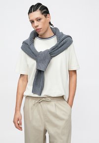 Pull gris en maille enroulé autour d'un T-shirt blanc, avec une texture côtelée et une coupe décontractée. Pantalon beige clair et coiffure soignée.