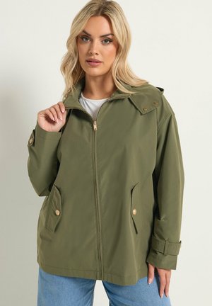 Vrouw draagt een olijfgroene jas met ritssluiting, met zakken met knopen en manchetten met riempjes, gecombineerd met lichtblauwe spijkerbroek, tegen een effen achtergrond.