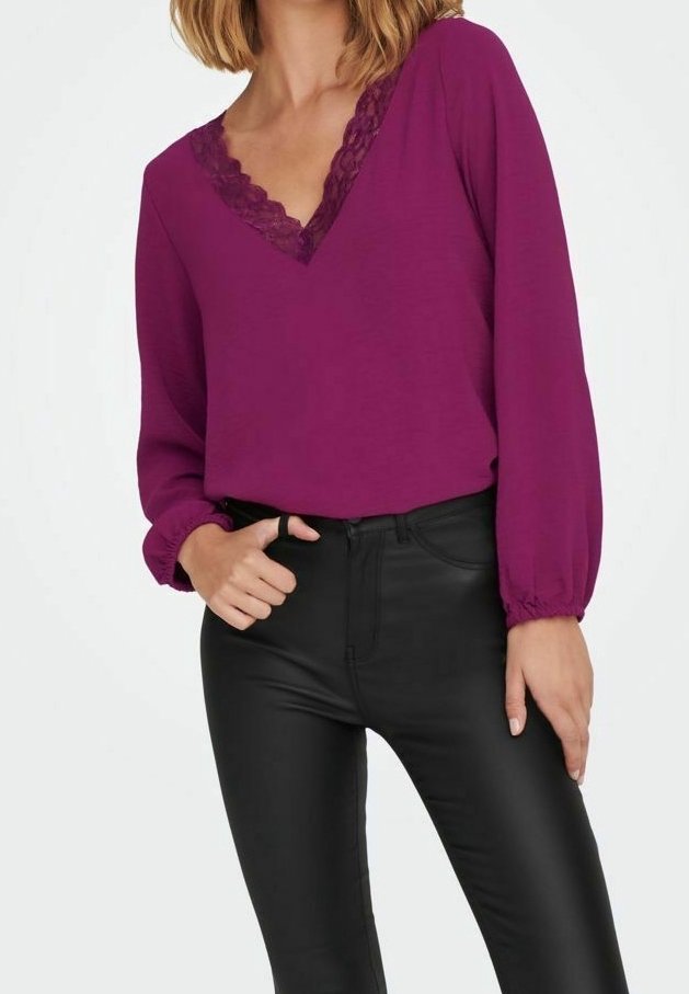 Femme portant un chemisier violet à manches longues avec col en V en dentelle et un pantalon noir ajusté, posant avec une main dans la poche et l’autre reposant sur la cuisse.