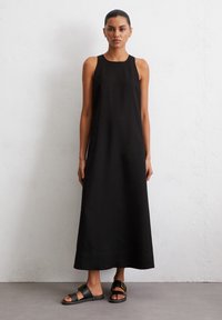 Abito lungo nero senza maniche con scollatura rotonda, realizzato in tessuto liscio. Indossato con sandali neri caratterizzati da un dettaglio con fibbia.