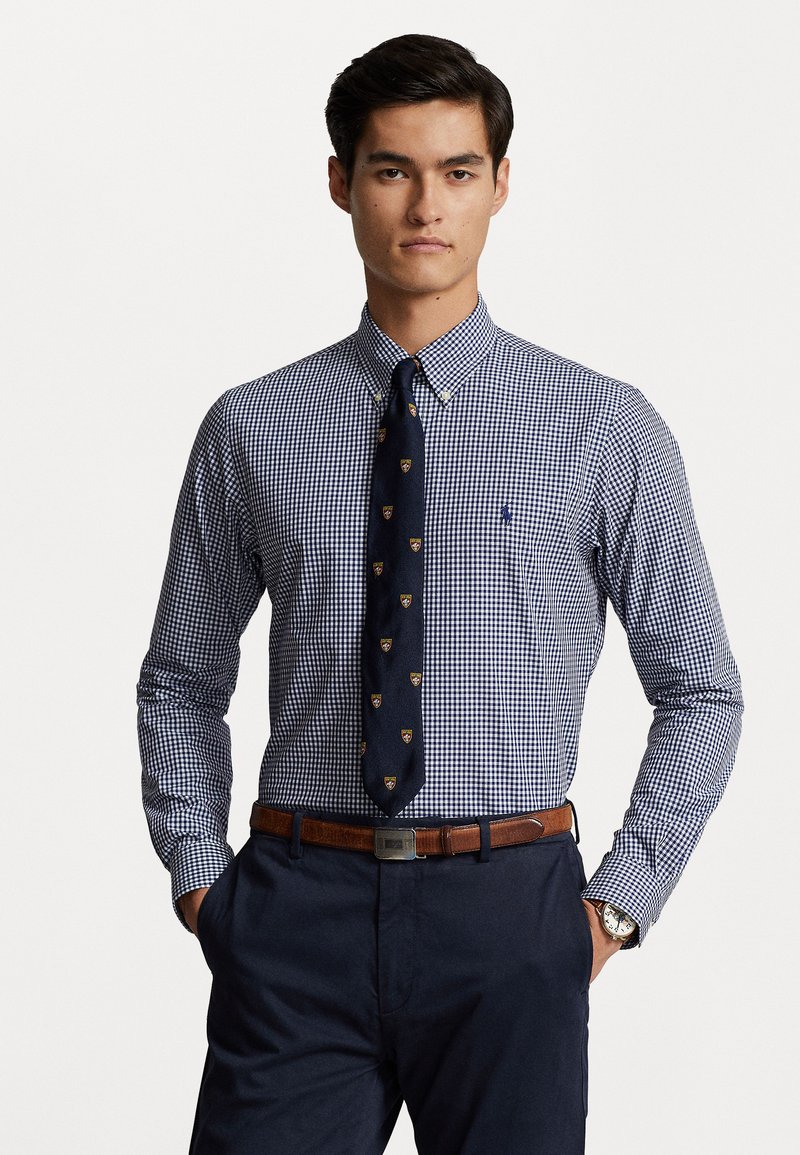 Chemise à manches longues à carreaux bleu marine et blancs avec col boutonné, associée à une cravate foncée ornée d'un motif et une ceinture marron.