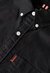 Camicia nera con bottoni bianchi, presenta una tasca con etichetta arancione "Superdry", materiale morbido e colletto cucito con precisione.