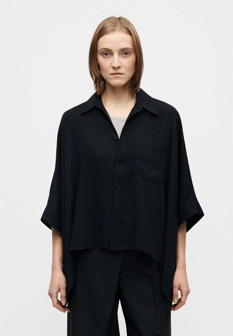 Chemise noire oversize à manches courtes, boutonnée sur le devant, avec une poche poitrine. Tissu léger à texture lisse.