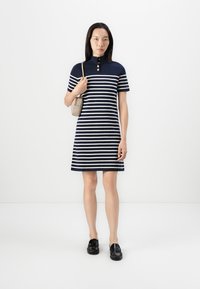 Navy og hvite stripete kortermet kjole med høy krage og tre knapper. Modellen holder en liten beige veske og har på seg svarte sko.