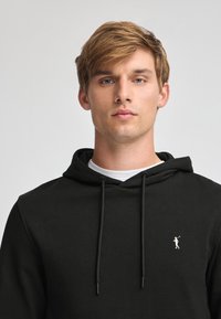 Felpa nera con cappuccio, dotata di tasca a marsupio frontale, cordini regolabili e un piccolo logo dorato sulla parte sinistra del petto.