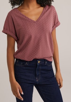 Woman wearing a mauve textured V-neck short-sleeve blouse tucked into dark blue denim jeans with rolled cuffs.