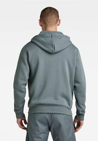 Felpa con cappuccio in teal smorzato, realizzata in morbido misto cotone. Polsini e orlo a coste, tasca a marsupio, cuciture piatte e una toppa con logo in vita.