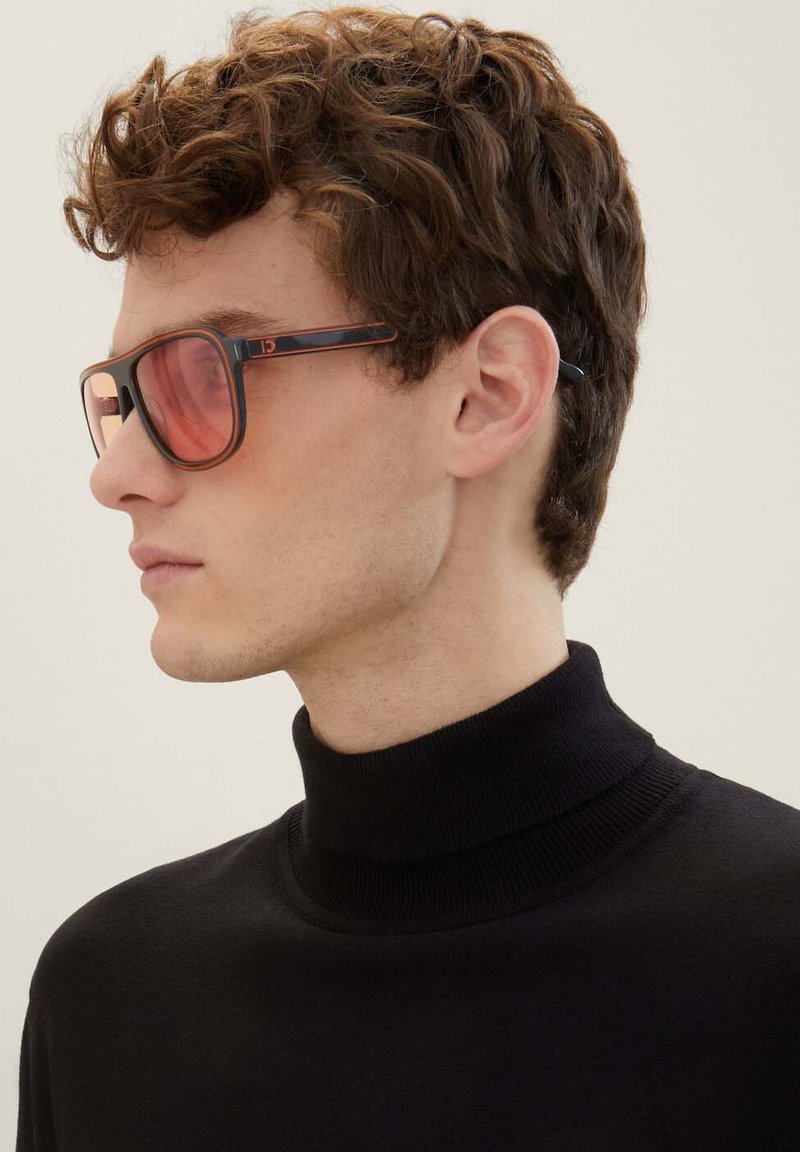 Okulary przeciwsłoneczne z ciemnym oprawą w kratę żółwia, różowymi soczewkami i kontrastującymi pomarańczowymi dodatkami. Modelka ma na sobie czarny sweter z golfem.