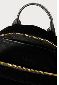 Melna teksturēta rokassoma ar noapaļotu rokturi un atvērtu zelta rāvējslēdzēju, kas rāda gludu melnu audumu iekšpusi ar izšūtu kabatu.