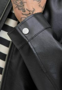 Puño de chaqueta de cuero negro con un botón de presión plateado, que muestra una textura suave y un primer plano de la tela a rayas de abajo.