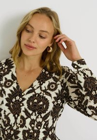 Robe à motif floral en crème et noir. Caractéristiques : manches longues, décolleté en V et détails de boutons. Accessoirisée avec de grandes boucles d'oreilles en or.