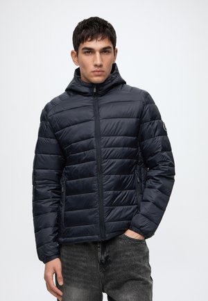 Jeune homme portant une veste matelassée noire à capuche avec des poches zippées et un jean gris foncé, debout contre un fond clair uni.