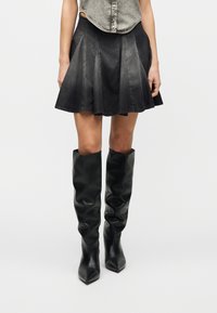 Γυναίκα που φορά φθαρμένη μαύρη τζιν φούστα και χαλαρά μαύρα ψηλοτάκουνα μποτάκια με μυτερή μύτη μέχρι τα γόνατα, σε απλό φόντο.