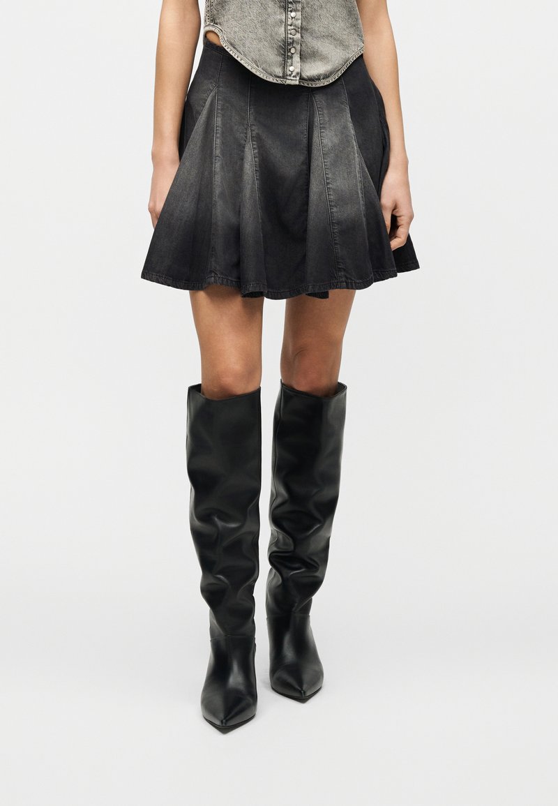 Γυναίκα που φορά φθαρμένη μαύρη τζιν φούστα και χαλαρά μαύρα ψηλοτάκουνα μποτάκια με μυτερή μύτη μέχρι τα γόνατα, σε απλό φόντο.