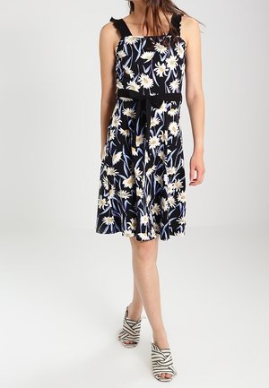 Vrouw draagt een zwarte mouwloze jurk met witte madeliefjesprint en een zwarte tailleband, gecombineerd met zwart-witte gestreepte openhakken.