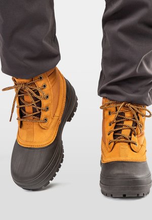 Persoon die bruine en zwarte waterdichte winterlaarzen met dikke zolen en een zwarte broek draagt.