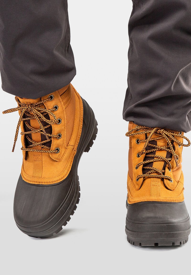 Personne portant des bottes d'hiver imperméables marron et noires avec des semelles épaisses et un pantalon noir.