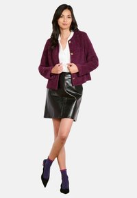 Femme portant une blouse blanche, un cardigan violet boutonné, une jupe en cuir noire, des chaussettes à carreaux violettes et des chaussures noires à bouts pointus, se tenant avec les jambes croisées.