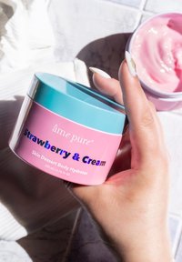 Rosa burk märkt "Jordgubb & Grädde" med ett ljusblått lock, innehållande gräddfuktighetskräm. Hand som håller burken med glansig yta.