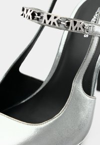 Ασημένιο μεταλλικό ψηλοτάκουνο παπούτσι με ανοιχτές πλευρές, που διαθέτει μαύρο πάτο και ασημένια αλυσίδα με λεπτομέρεια λογότυπου στο λουράκι.