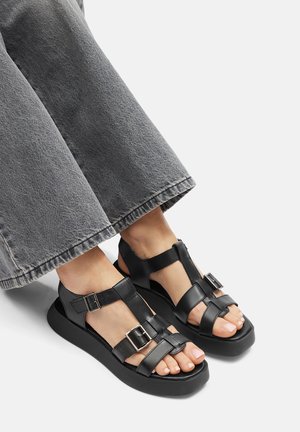 Schwarze Ledersandalen mit flacher Sohle und verstellbaren Schnallen, mit offener Zehenpartie und T-Riemen-Design. Kombiniert mit grauen Hosen mit weitem Bein.