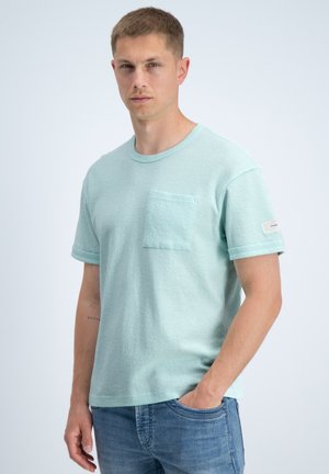 Jonge man met een lichtblauw kort mouwhemd met borstzak en blauwe spijkerbroek, staand met één hand in de zak tegen een effen achtergrond.