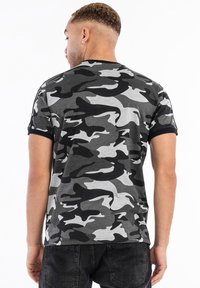 Pilka kamufliažo t-shirt su juodais akcentais, trumpomis rankovėmis ir apvaliu kaklu. Pagamintas iš medvilnės su lygiu tekstūra. Kasdienis sijonas.