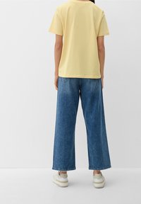 Camiseta de algodón amarilla con mangas cortas, combinada con culottes de mezclilla azules de corte holgado. Zapatillas visibles en la parte inferior. Diseño simple y ajuste relajado.