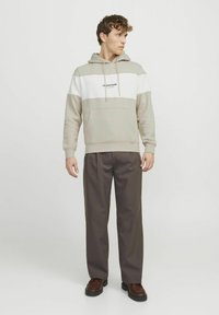 Herre sweatshirt i beige og hvid, med en frontlomme, snor i hætten og broderet logo. Sættet er kombineret med mørkebrun bukser og sko.